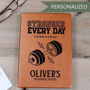 May include: A brown leather fitness journal with the text "STRONGER EVERY DAY FITNESS JOURNAL" and "OLIVER'S TRAINING NOTES". A barbell graphic is centered below the text. The word "PERSONALIZED" is in a green banner at the top.