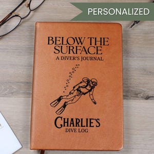 Può includere: Un diario in pelle marrone con il testo "BELOW THE SURFACE A DIVER'S JOURNAL" e "CHARLIE'S DIVE LOG". Un'illustrazione di un subacqueo è presente sulla copertina. La parola "PERSONALIZZATO" è in alto. Occhiali e penna visibili.