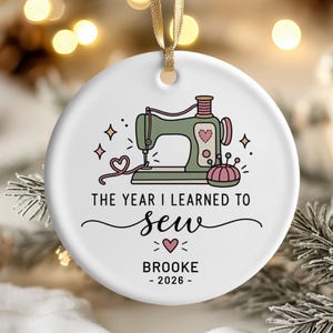 Könnte beinhalten: Ein rundes, weißes Keramikornament mit einem goldenen Band zum Aufhängen. Das Design zeigt eine grüne Nähmaschine, ein rosafarbenes Nadelkissen und Herzen. Der Text lautet: THE YEAR I LEARNED TO sew, BROOKE und 2026, ergänzt durch kleine Sterne.