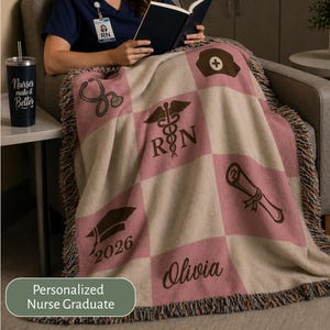 May include: A personalized nurse graduate blanket with a pink and beige checkered pattern. The blanket features medical symbols, a graduation cap with the year 2026, and the name Olivia. A black tumbler with the text "Nurses make it Better" is on a side table.