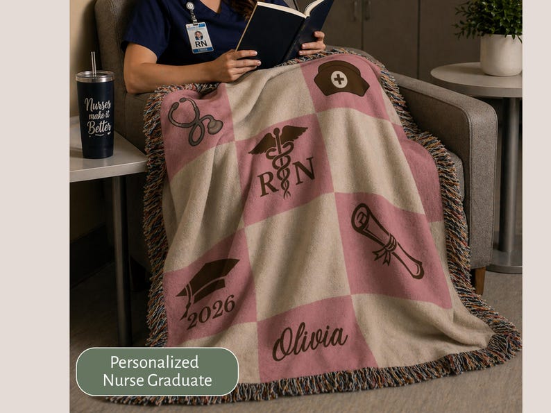 May include: A personalized nurse graduate blanket with a pink and beige checkered pattern. The blanket features medical symbols, a graduation cap with the year 2026, and the name Olivia. A black tumbler with the text "Nurses make it Better" is on a side table.
