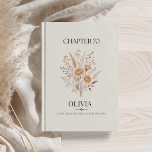 May include: A hardcover book with a cream-colored cover featuring the text CHAPTER 70 at the top, a central illustration of a dried floral bouquet in beige and brown tones, the name OLIVIA in the center, and the text EVERY CHAPTER BRINGS NEW GROWTH.
