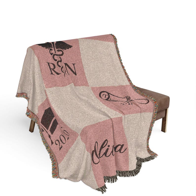 May include: A woven throw blanket with a patchwork design in pink and beige. The blanket features a graduation cap, the text "2020", a caduceus symbol with "RN", and the name "Olivia". The blanket has a fringed edge.