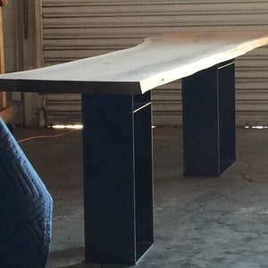May include: A long, live edge wooden table with a natural wood finish. The table is supported by two black metal legs.