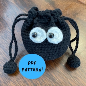 May include: A black crocheted drawstring bag with large white eyes and black pupils. The bag has a round shape and is decorated with a pair of white, crocheted eyes with black centers. The text "PDF PATTERN!" is in a blue circle.