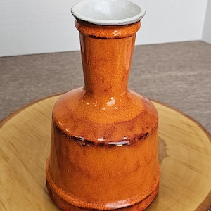May include: A small, orange ceramic vase with a white rim. The vase has a rounded base and a narrow neck.