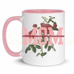 May include: White ceramic mug with a pink rim and handle. The mug features a floral design with pink roses and green leaves. The text "MOM" is printed in pink on the mug with the text "TO THE WORLD, YOU ARE A MOTHER. TO ME, YOU ARE THE WORLD" below it.