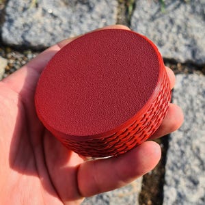 Op de afbeelding: Een rond, rood object met een getextureerd oppervlak en een gevlochten zijkant. Het object wordt in een hand gehouden. De achtergrond bestaat uit grijze stenen. De rode kleur is uniform.