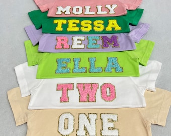 May include: A stack of six t-shirts with different names and numbers on them. The shirts are in various colors, including pink, green, purple, and white. The names and numbers are made of glitter and have a gold outline. The names are Molly, Tessa, Reem, Ella, Two, and One.
