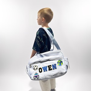 May include: A light blue shoulder bag with a zipper closure and a light blue strap. The bag has the name "OWEN" in blue and black letters, along with patches of a soccer ball, a police car, and a globe.