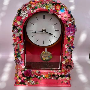 May include: A pink arched mantel clock featuring a white clock face with black hands and no numerals. The frame is heavily decorated with a dense arrangement of colourful miniature plastic flowers, leaves, and small bee figurines. A round pendulum hangs inside.