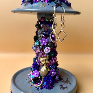 May include: A gray, decorative earring holder with a round base and a mushroom-shaped top. The holder is adorned with purple and blue sequins, flowers, and a gold cat figurine. Silver earrings hang from the top.