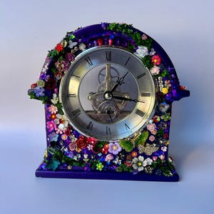 May include: A purple arched mantel clock featuring a silver face with Roman numerals and visible internal gears. The frame is decorated with an array of colourful miniature flowers, green foliage, small butterflies, and sparkling glitter accents.