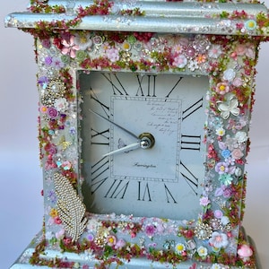 May include: A decorative clock with a silver frame, embellished with colourful artificial flowers, butterflies, and decorative elements. The clock face displays Roman numerals and the name "Farrington". The clock is approximately 15 cm wide and 20 cm tall.