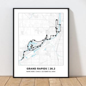May include: Framed print featuring a black line route map of Grand Rapids, Michigan, with the text "GRAND RAPIDS | 26.2". The map includes a route marked with black dots, and the text "NAME HERE | 3:48:12 | OCTOBER XX, XXXX". The print is in a black frame.