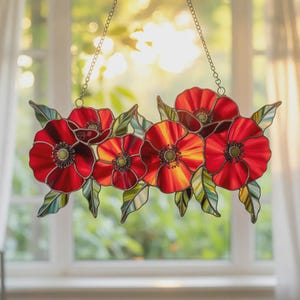 May include: A stained glass window hanging featuring six red poppies with yellow and orange centers and green leaves. The poppies are arranged horizontally and suspended by a chain. The window is in front of a window with sunlight shining through.