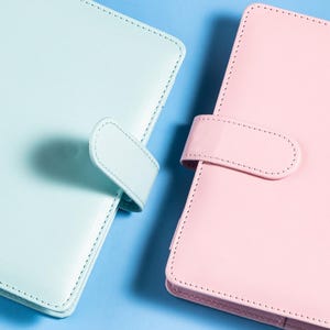 Puede incluir: Primer plano de dos organizadores encuadernados en cuero, uno en azul pastel suave y otro en rosa delicado. Cada uno tiene un cierre de lengüeta a juego asegurado con un diseño cosido. El fondo es azul claro liso.