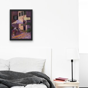 May include: Framed painting of a European street scene. The artwork depicts a building with a white awning, a sign, and a few figures at tables. The color palette includes shades of purple, yellow, and green.