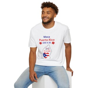 Puede incluir: Camiseta blanca con el texto "Moca Puerto Rico" en azul y rojo, junto con los números "100 X 35" y detalles florales. Debajo, un gráfico del mapa de Puerto Rico con la bandera y el texto "En mi ADN".