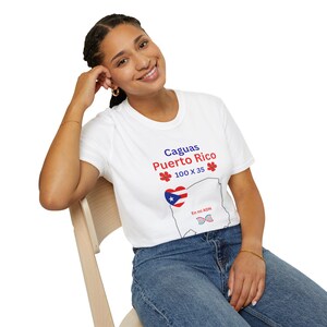 Puede incluir: Camiseta blanca con el texto "Caguas Puerto Rico" en rojo y azul, junto con un contorno del mapa de Puerto Rico y la bandera puertorriqueña. La camiseta también presenta el texto "100 X 35" y "En mi ADN".