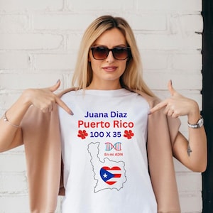 Puede incluir: Camiseta blanca con el texto "Juana Diaz Puerto Rico" en azul y rojo, junto con un gráfico de la bandera de Puerto Rico dentro de un corazón. La camiseta también presenta el texto "100 X 35" y "En mi ADN".