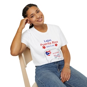Puede incluir: Camiseta blanca con el texto "Lajas Puerto Rico 100 x 35" en azul y rojo. La camiseta presenta un mapa de Puerto Rico con la bandera y las palabras "En mi ADN" con un gráfico de hebra de ADN.