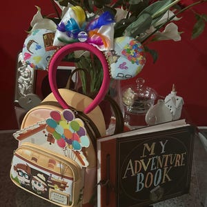 Può includere: Uno zaino giallo e marrone con una casa e palloncini su di esso. Lo zaino è accanto a un libro marrone con le parole "My Adventure Book" sulla copertina. Ci sono anche orecchie di Minnie Mouse rosa e blu con un fiocco arcobaleno in cima.