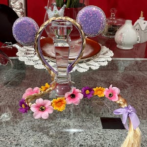 Pode incluir: Faixa de cabeça roxa e dourada com uma coroa, duas orelhas roxas e uma longa trança loira com flores rosa e amarelas.