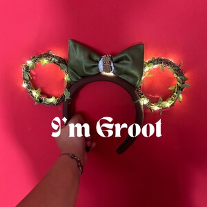 May include: A brown headband with green bow and Groot character embellishment. The headband features two circular ears decorated with faux vines and fairy lights. The text "I'm Groot" is displayed in white script.