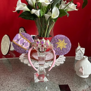 Puede incluir: Orejas de Minnie Mouse moradas y doradas con una corona y un diseño de sol. Las orejas están en una diadema rosa y se muestran en un soporte transparente.