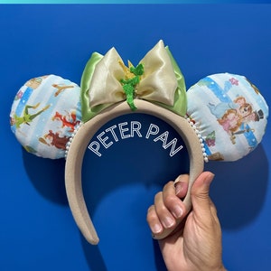 Può includere: Orecchie di Minnie Mouse a tema Peter Pan con un fiocco verde e giallo, con una figura verde di Peter Pan sul fiocco. Le orecchie sono ricoperte da una stampa di tessuto con Peter Pan, Trilly e altri personaggi del film.