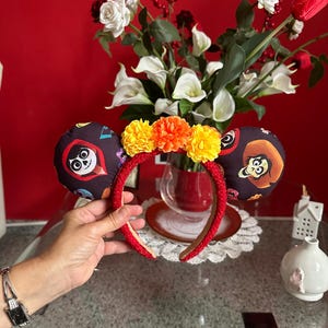 Può includere: Fascia rossa con orecchie in tessuto nero con personaggi del film Coco. Le orecchie sono decorate con fiori di tagete gialli e arancioni.