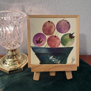 May include: A watercolor painting of a bowl of fruit in a dark blue bowl, displayed on a small wooden easel. The fruit is depicted in shades of purple, red, and green. The painting is framed in a light wood frame.