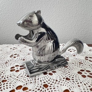 May include: A polished silver-toned metal nutcracker shaped like a squirrel. The squirrel is depicted in a seated position on a rectangular base, holding a small object between its front paws, with a curved tail extending behind its body.