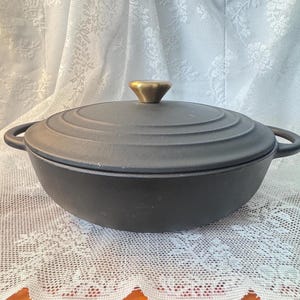 May include: A matte black cast iron Dutch oven with a round, low-profile body and two side handles. The matching lid features concentric circular ridges and a central, conical-shaped metallic gold knob for lifting the heavy cookware.
