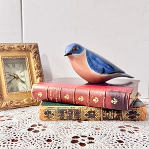 May include: A ceramic bluebird figurine with a blue back and peach-colored chest, perched atop two stacked vintage books. The top book is red with gold lettering, and the bottom book is green with gold details, next to a gold-framed clock.