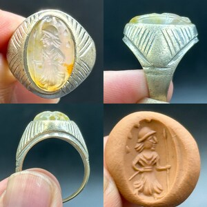 May include: A silver ring with a clear agate stone set in a bezel. The stone features a carved image of a woman in a helmet holding a spear. The ring is shown from multiple angles, including a close-up of the stone and a view of the ring on a finger.
