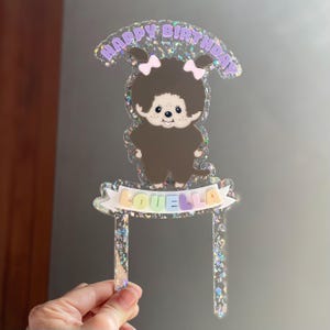 May include: A clear acrylic cake topper with a cartoon character and the text "HAPPY BIRTHDAY" in purple. The character has brown fur, pink bows, and the name "LOUELLA" on a banner.