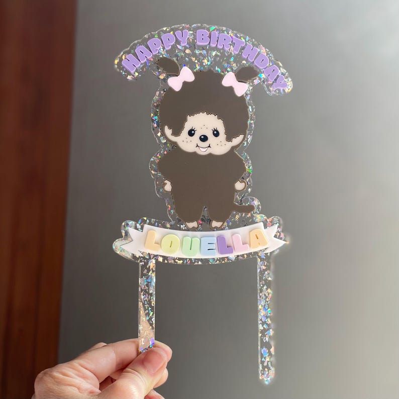 May include: A clear acrylic cake topper with a cartoon character and the text "HAPPY BIRTHDAY" in purple. The character has brown fur, pink bows, and the name "LOUELLA" on a banner.