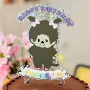 May include: A cake topper featuring a cartoon character with a dark brown body, a fluffy hairstyle, and two white bows. The topper has the words "HAPPY BIRTHDAY" and "LOVELLA" in a pastel color scheme.