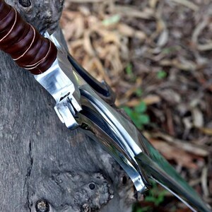 Peut inclure: Gros plan d'une &eacute;p&eacute;e avec une lame en acier poli et un manche envelopp&eacute; de cuir marron. L'&eacute;p&eacute;e repose sur une surface en bois vieilli, avec un arri&egrave;re-plan flou de feuilles et de brindilles.