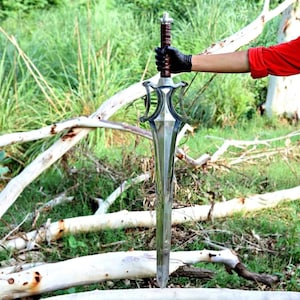 Peut inclure: Une grande &eacute;p&eacute;e argent&eacute;e avec une garde d&eacute;corative et un manche en cuir marron. La lame longue et pointue est mise en valeur sur un fond de feuillage vert et de branches d'arbres tomb&eacute;es. La longueur totale de l'&eacute;p&eacute;e est d'environ 90 cm.