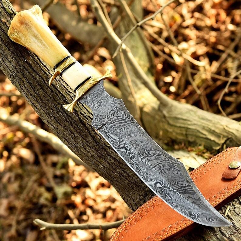 May include: A Damascus steel hunting knife with a bone handle and brass accents. The blade has a distinctive pattern, and the knife rests on a tree branch with a leather sheath visible.