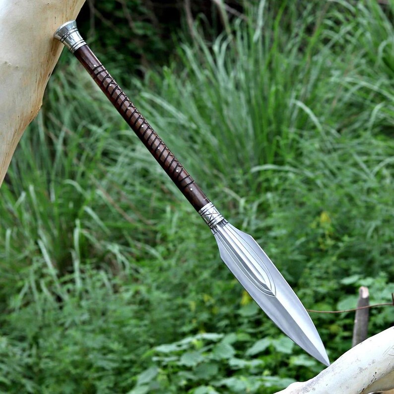 May include: A spear with a silver blade and a dark brown wooden shaft. The shaft is wrapped with a lighter brown material and has silver accents at the top and bottom. The spear is set against a backdrop of green foliage.