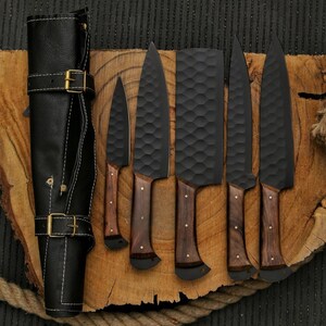 May include: A set of six black kitchen knives with wooden handles and a black leather knife roll. The blades have a textured surface. The knife roll is closed with two buckles.