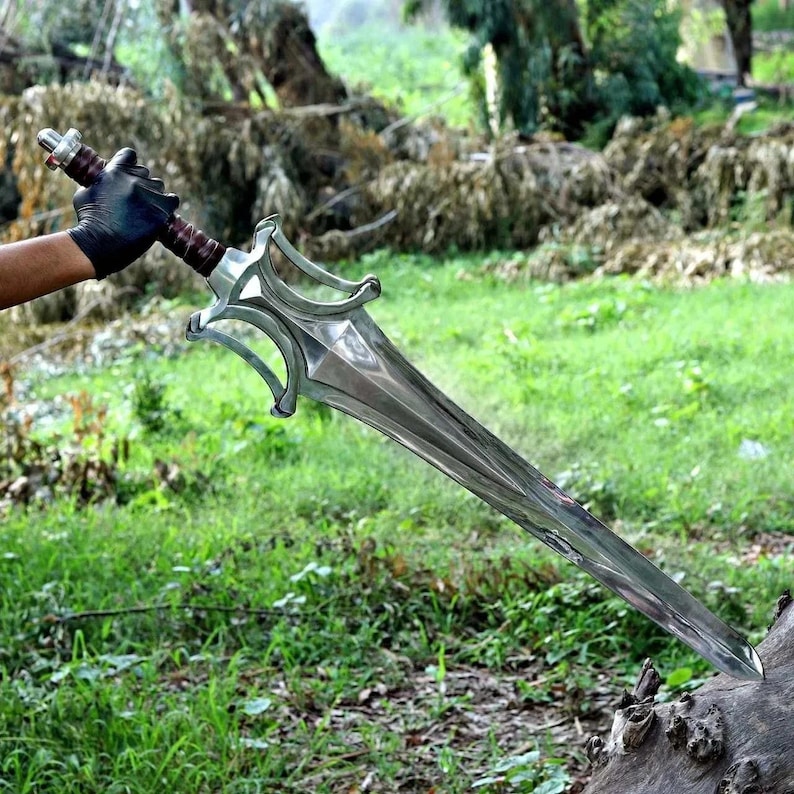 Peut inclure: Une &eacute;p&eacute;e argent&eacute;e avec une longue lame pointue et une poign&eacute;e orn&eacute;e. La poign&eacute;e a une poign&eacute;e marron et une garde d&eacute;corative. L'&eacute;p&eacute;e est tenue sur fond d'herbe verte et de feuillage.