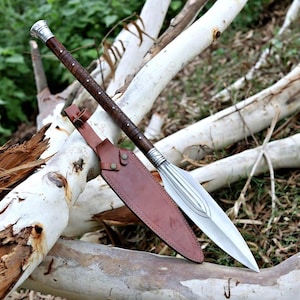 May include: A spear with a polished silver blade and a dark brown wooden shaft wrapped in leather. The spear is accompanied by a brown leather sheath. The spear is resting on a white tree branch.