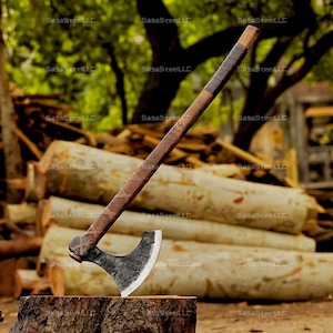 Könnte beinhalten: Eine schwarze und braune Axt mit einem Holzstiel und Lederumwicklungen. Die Axt liegt auf einem Baumstumpf mit einem Holzstapel im Hintergrund.