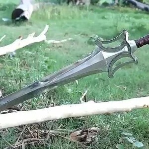 Peut inclure: Une &eacute;p&eacute;e argent&eacute;e avec une garde d&eacute;corative et un manche en cuir marron. L'&eacute;p&eacute;e est tenue par une main gant&eacute;e sur un fond d'herbe verte et de branches tomb&eacute;es.