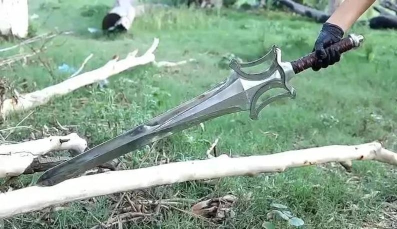 Peut inclure: Une &eacute;p&eacute;e argent&eacute;e avec une garde d&eacute;corative et un manche en cuir marron. L'&eacute;p&eacute;e est tenue par une main gant&eacute;e sur un fond d'herbe verte et de branches tomb&eacute;es.